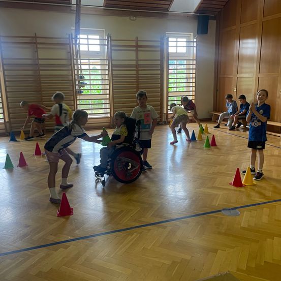 In einer Turnhalle spielen Kinder ein Spiel mit Kegel. Einige Kinder stehen, andere sind im Rollstuhl. Einige klatschen.