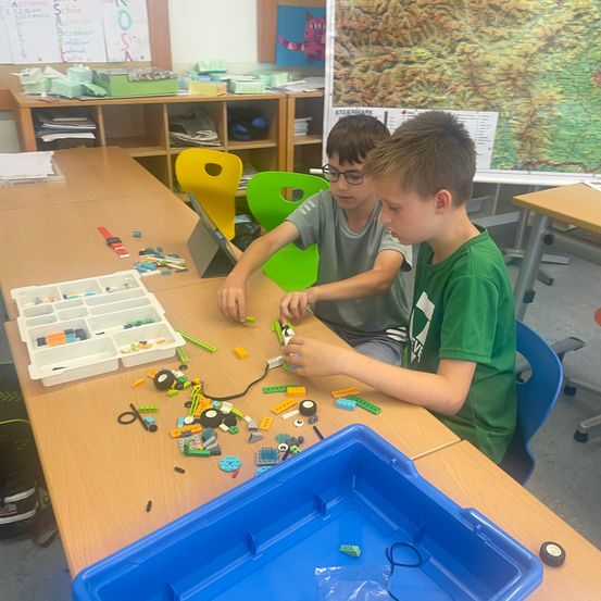 Zwei Jungen sitzen an einem Tisch und arbeiten an einem Projekt mit Lego-Steinen. Ein Junge trägt eine Brille und der andere ein grünes T-Shirt.