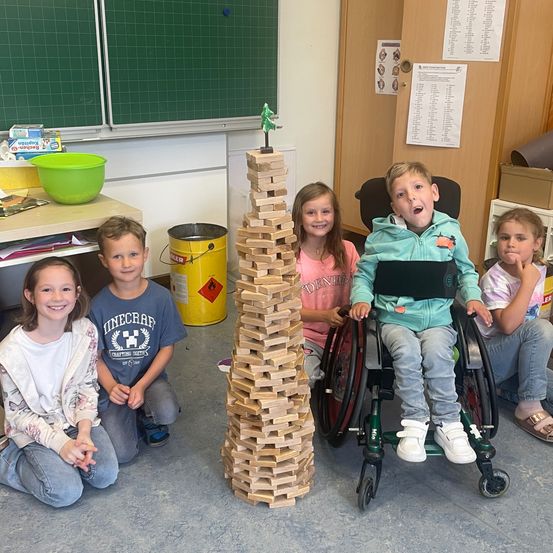 Fünf Kinder sitzen um einen hohen Holzturm herum auf dem Boden. Ein Kind im Rollstuhl. Dahinter ein grüner Eimer und ein gelber Mülleimer.