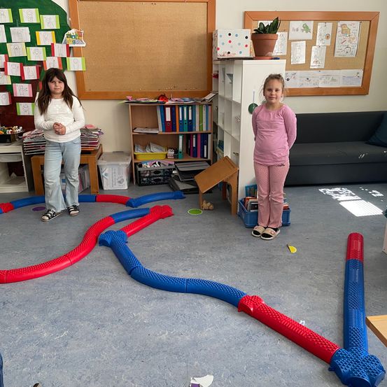 Zwei junge Mädchen stehen in einem Klassenzimmer mit einem Labyrinth auf dem Boden. Ein Mädchen sieht ernst aus, während das andere zur Seite schaut. Dahinter ist eine Pinnwand mit Zeichnungen beheftet.