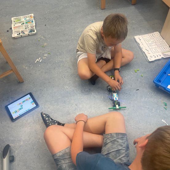 Zwei Jungen bauen Legosteine auf dem Boden, einer sitzt und der andere liegt. Ein Tablet mit bunten Quadraten steht neben ihnen.