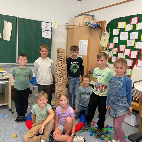 Mehrere Kinder stehen um einen Turm aus Blöcken in einem Klassenzimmer. Ein Junge sitzt auf einem roten Ball.