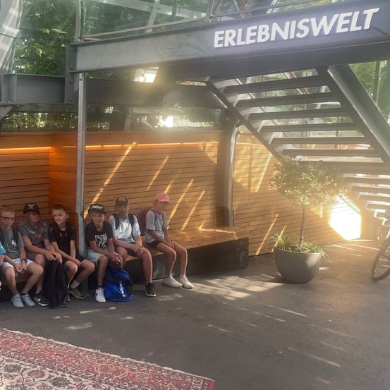 Eine Gruppe junger Leute sitzt auf einer Bank vor einem Gebäude mit einem Schild, das 'Erlebniswelt' steht. Sie tragen Mützen und Turnschuhe. Ein Topf mit Pflanzen und ein Fahrrad sind in der Nähe.
