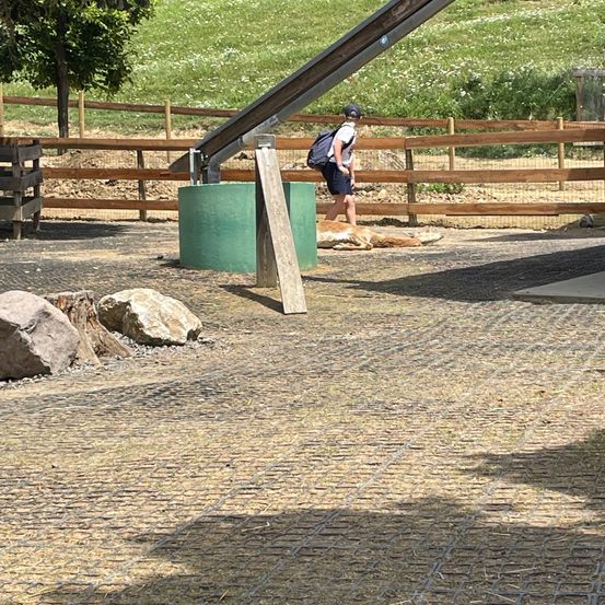Eine Person mit Rucksack geht an einem Zaun und einem großen grünen Fass vorbei. Ein Hund liegt in der Nähe auf dem Boden.