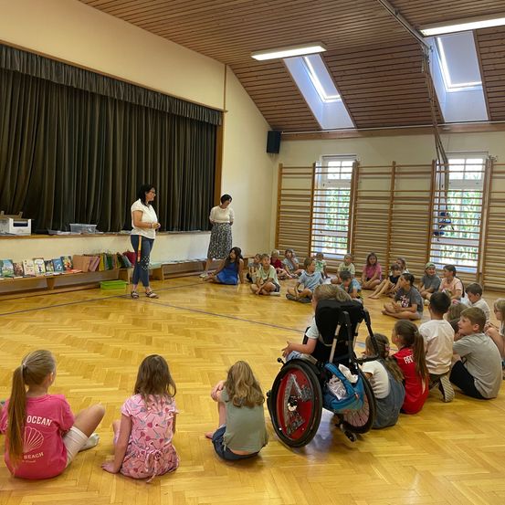 Eine Gruppe von Kindern sitzt im Kreis auf einem Holzboden und hört einer Frau zu, die vor ihnen steht. Der Raum hat Holzbalken und Fenster.