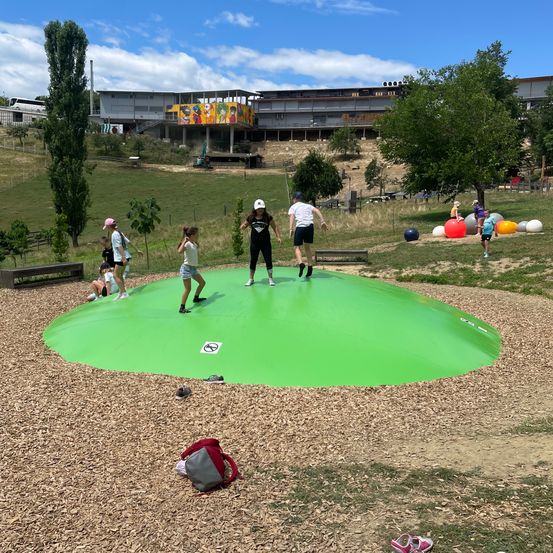 Eine Gruppe von Menschen spielt auf einem großen grünen aufblasbaren Pool im Freien. Einige stehen, während andere auf dem Gras sitzen. Im Hintergrund befinden sich bunte Bälle und ein Gebäude.