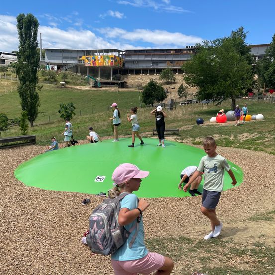 Kinder spielen auf einem großen grünen aufblasbaren Pool in einem Park mit einem Gebäude und Bäumen im Hintergrund.