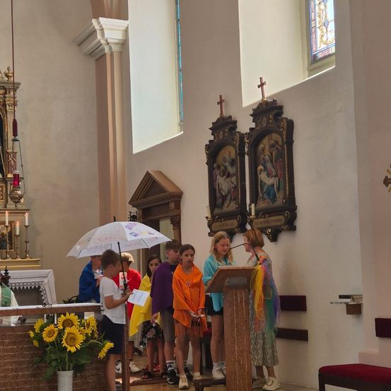 Eine Gruppe von Kindern steht in einer Kirche, einer hält einen Regenschirm und ein anderer spricht an einem Podium. Auf dem Altar stehen Sonnenblumen.