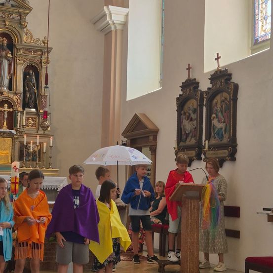 Eine Gruppe von Kindern in bunten Umhängen steht in einer Kirche, einer hält einen Regenschirm und ein anderer spricht an einem Podium.