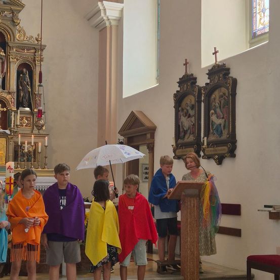 Eine Gruppe von Kindern in bunten Umhängen steht in einer Kirche, mit einer Frau, die an einem Podium spricht. Hinter ihnen befindet sich ein Altar mit Kerzen und religiösen Ikonen.