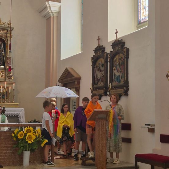Eine Gruppe von Kindern und Erwachsenen steht in einer Kirche, wobei eine Frau ein Mikrofon am Podium hält. Sonnenblumen und religiöse Symbole schmücken den Raum.