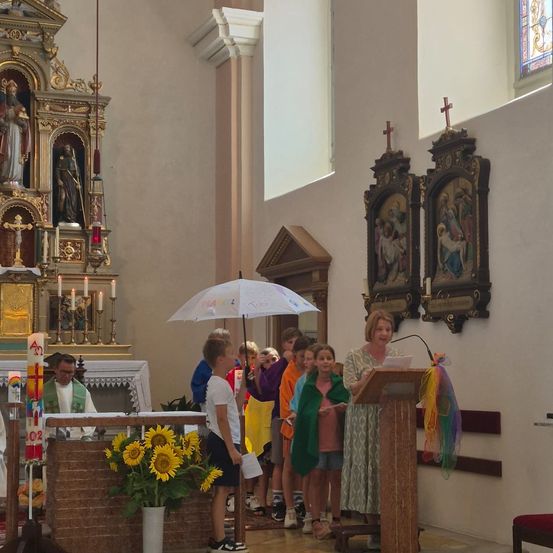 Eine Frau steht in einer Kirche an einem Podium und hält ein Papier in der Hand, während sich Kinder und ein Priester um sie herum versammeln. Im Vordergrund stehen Sonnenblumen auf einem Tisch.