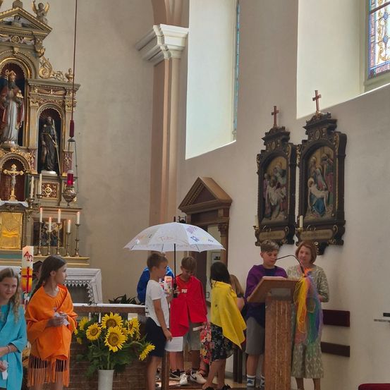 Eine Gruppe von Kindern und Erwachsenen steht in einer Kirche, einige halten Regenschirme. Eine Frau spricht an einem Podium, mit Sonnenblumen in einer Vase in der Nähe. Religiöse Ikonen schmücken die Wände.