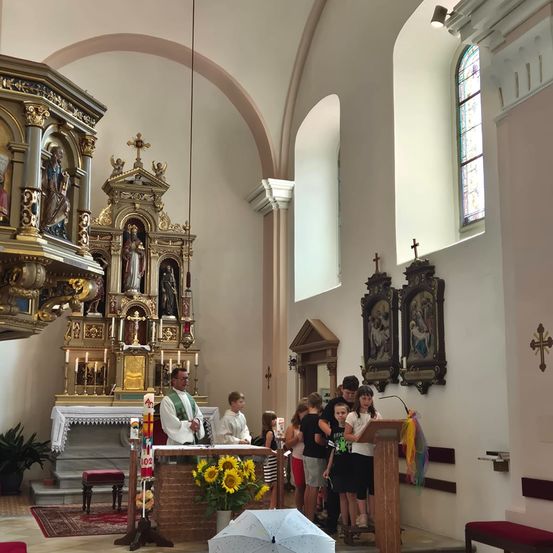 Ein Priester steht am Altar in einer Kirche, umringt von Kindern, die sich um ein Podium versammelt haben. Der Altar ist mit Kerzen und einer goldenen Statue geschmückt. Die Kirchenwände sind mit religiösen Gemälden und Buntglasfenstern verziert.