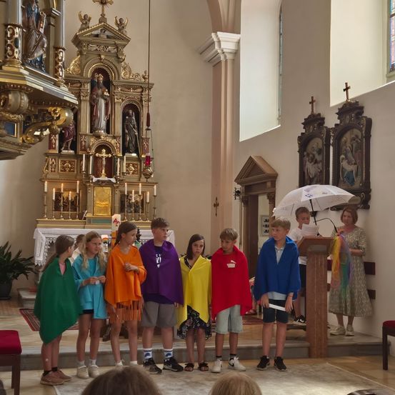 Eine Gruppe von Kindern in bunten Umhängen steht in einer Kirche, mit einem Altar und religiösen Statuen im Hintergrund.
