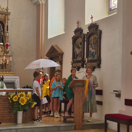 Eine Gruppe von Kindern und einer Frau stehen in einer Kirche, die Frau hält ein Mikrofon an einem Podium. Sonnenblumen stehen auf einem Tisch links.