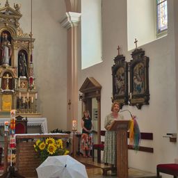 Eine Frau steht an einem Podest in einer Kirche, mit einem Strauß Sonnenblumen und einem weißen Regenschirm in der Nähe. Eine andere Frau steht hinter ihr, und religiöse Ikonen schmücken die Wände.
