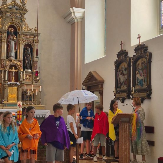 Eine Gruppe von Kindern in bunten Umhängen steht in einer Kirche, während eine Frau ein Mikrofon an einem Rednerpult hält. Dahinter steht ein Priester an einem Altar mit Kerzen.