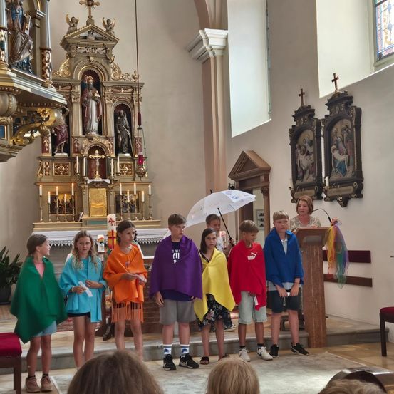 Eine Gruppe von Kindern in bunten Umhängen steht in einer Kirche, mit einer Frau, die einen Regenschirm hinter sich hält.
