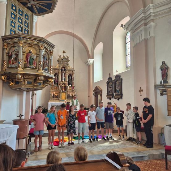 Eine Gruppe von Kindern in einer Kirche, die vor einem Altar mit einem Priester stehen. Sie sind in verschiedenen Farben gekleidet und scheinen an einer Zeremonie teilzunehmen.