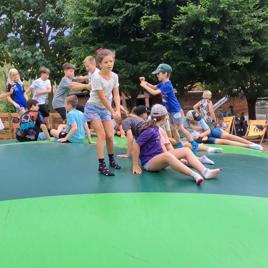 Eine Gruppe von Kindern spielt auf einer großen aufblasbaren Matte im Freien. Einige stehen, während andere sitzen oder liegen. Bäume und eine Ziegelmauer sind im Hintergrund.