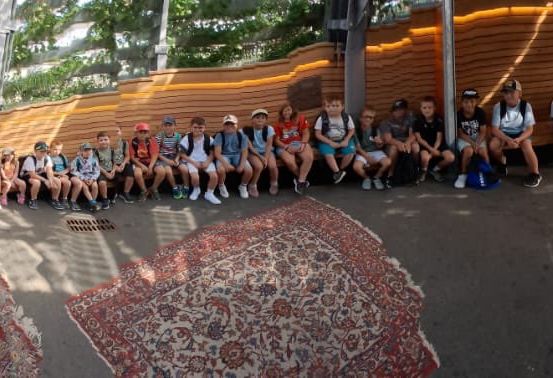 Eine Gruppe von Kindern und Erwachsenen sitzt auf einer Bank im Freien, mit einem Holzzaun und einem Teppich auf dem Boden. Sie tragen alle Freizeitkleidung.