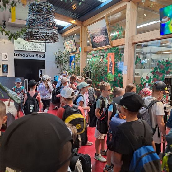 Eine Gruppe von Kindern mit Rucksäcken wartet in einer Schlange am Museumseingang. Der Eingang ist mit 'Laboko-Saal' beschriftet. Es gibt Pflanzen und Kunstwerke an den Wänden.