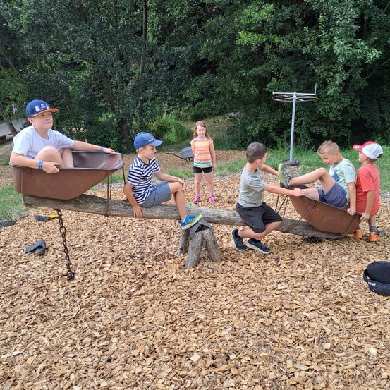 Fünf Kinder spielen auf einer Wippe in einem Park. Ein Kind sitzt auf einer Holzplanke, während die anderen auf einer rostigen Schubkarre sitzen.