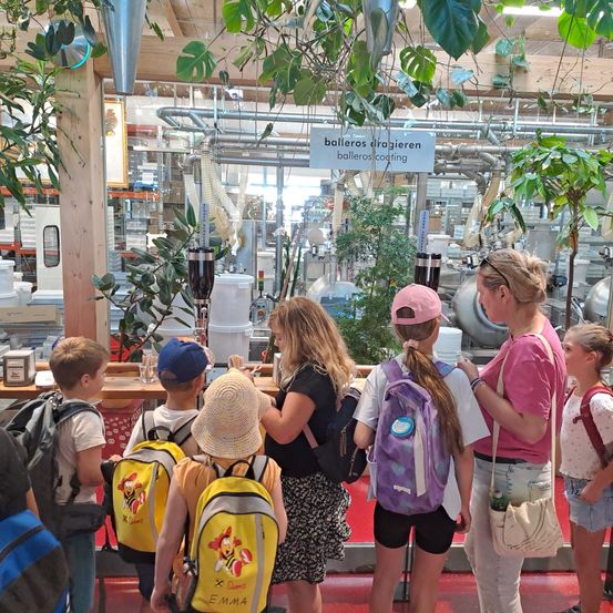 Eine Gruppe von Kindern und Erwachsenen steht in einer Fabrik mit Pflanzen und Rohren. Die Kinder tragen Rucksäcke und Hüte. Die Erwachsenen tragen Sonnenbrillen und Uhren. Im Hintergrund befindet sich ein Schild mit der Aufschrift 'balleros dragieren balleros cooling.'