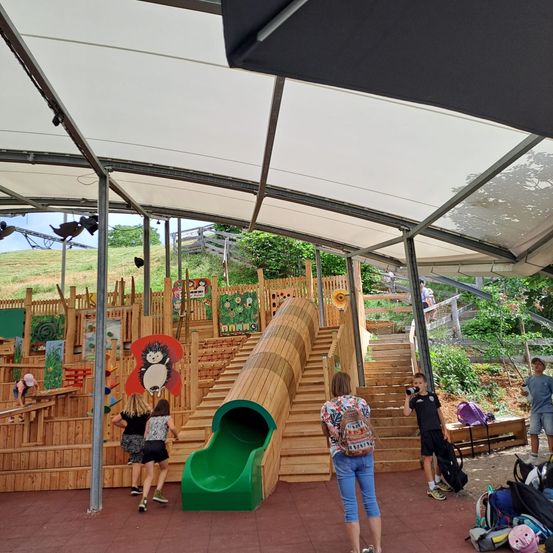 Eine Gruppe von Kindern und Erwachsenen spielt in einem hölzernen Spielbereich mit einer grünen Rutsche unter einem weißen Vordach.