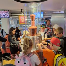 Eine Gruppe von Kindern und Erwachsenen versammelt sich um eine Schokoladenfontäne in einem Museum, einige tragen Hüte und Brillen.