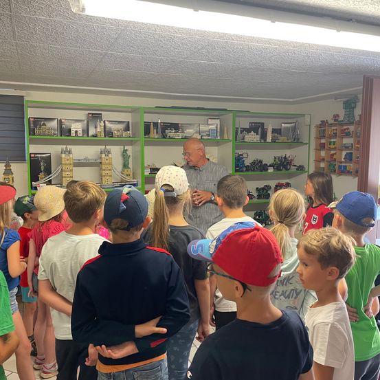 Eine Gruppe von Kindern steht in einem Raum und sieht einem Mann zu, der zu ihnen spricht. Der Raum hat Regale, die mit verschiedenen Gegenständen gefüllt sind, darunter Spielzeug und Bücher. Die Kinder tragen Mützen und Turnschuhe.