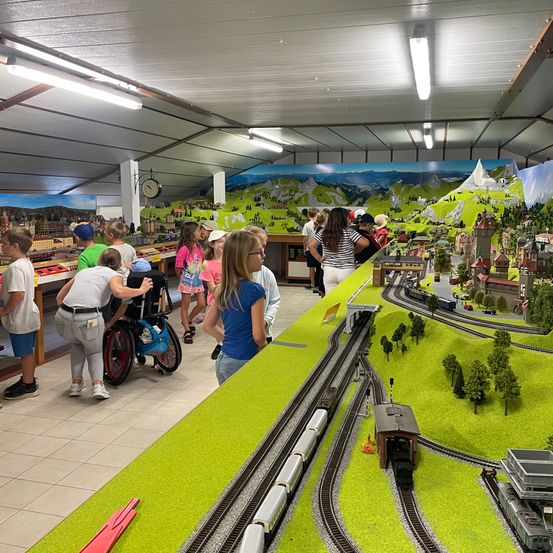 Eine Gruppe von Kindern und Erwachsenen versammelt sich um eine große Modelleisenbahn in einem Raum mit einer Wandmalerei von Bergen und einem Dorf. Die Kinder stehen und beobachten die Züge, während einige Erwachsene herumlaufen.