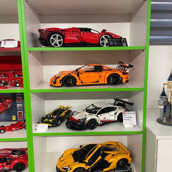 Eine Ausstellung von vier Lego-Sportwagen auf Regalen, darunter ein roter Ferrari, ein orangefarbener McLaren, ein weißer Porsche und ein gelber McLaren.