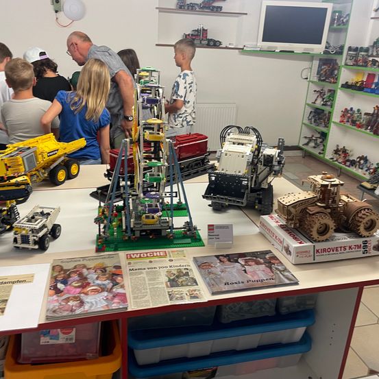 Eine Gruppe von Kindern und Erwachsenen versammelt sich um einen Tisch, auf dem verschiedene Lego-Kreationen ausgestellt sind, darunter ein Traktor, ein Riesenrad und ein Baufahrzeug. Der Raum ist mit Spielzeugregalen gefüllt und an der Wand befindet sich ein Monitor.