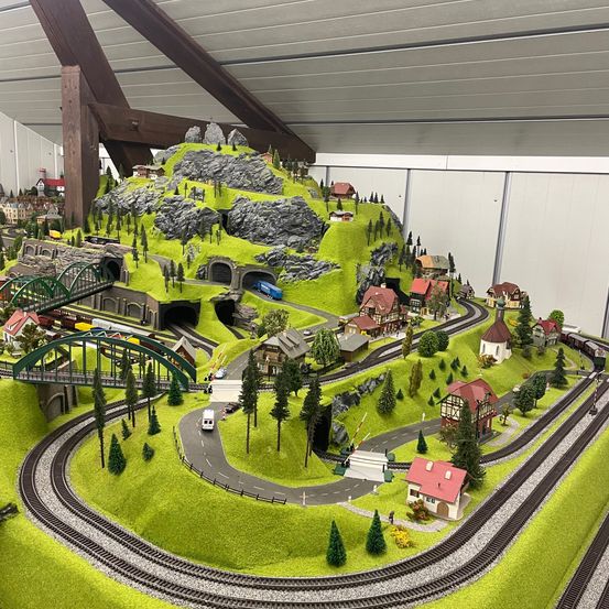Ein detailliertes Modell einer bergigen Region mit einer Eisenbahn, Häusern und einer Brücke. Die Szene umfasst einen Tunnel mit einfahrendem Zug und eine Straße mit Autos und Fußgängern.