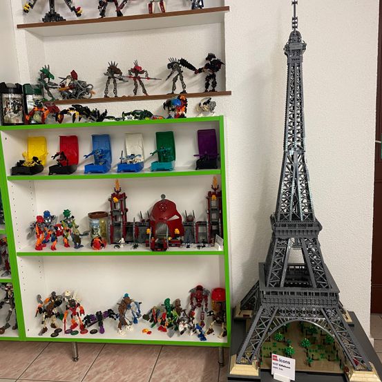 Ein Raum mit einer weißen Wand und einem gefliesten Boden. Ein Regal mit verschiedenen Lego-Sets, einschließlich eines Lego-Eiffelturms, steht links. Der Lego-Eiffelturm steht auf einer schwarzen Basis.