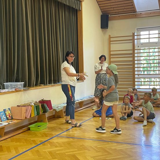 Eine Frau in einem Klassenzimmer verteilt Papieren an ein junges Mädchen, während andere Kinder auf dem Boden sitzen.