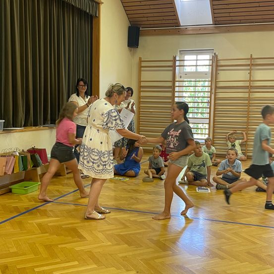 Eine Frau überreicht einem Mädchen in einem Klassenzimmer eine Urkunde, während andere Kinder auf dem Boden sitzen.