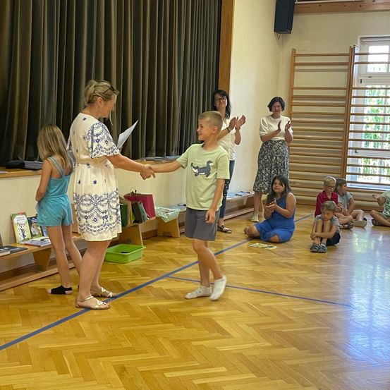 Eine Frau überreicht einem jungen Jungen ein Zertifikat in einem Raum, in dem andere Kinder sitzen und stehen.
