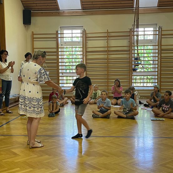 Eine Frau steht in einer Turnhalle und klatscht für einen jungen Jungen, der über den Boden geht. Mehrere Kinder sitzen auf dem Boden und schauen zu.