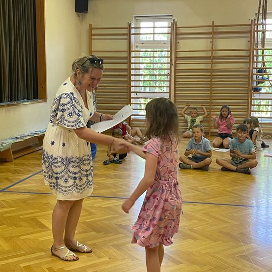 Eine Frau in einem weißen Kleid überreicht einer kleinen Mädchen in einem rosa Kleid eine Urkunde in einem Klassenzimmer. Mehrere Kinder sitzen hinter ihnen auf dem Boden.