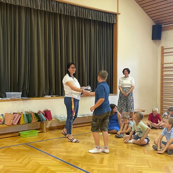 Eine Frau überreicht einem Jungen eine Trophäe, während eine andere Frau hinter ihnen steht. Kinder sitzen auf dem Boden und schauen zu.