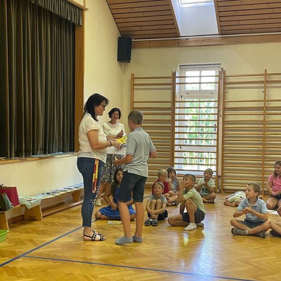 Eine Frau gibt einem Jungen ein Buch in einem Klassenzimmer, in dem andere Kinder auf dem Boden sitzen.