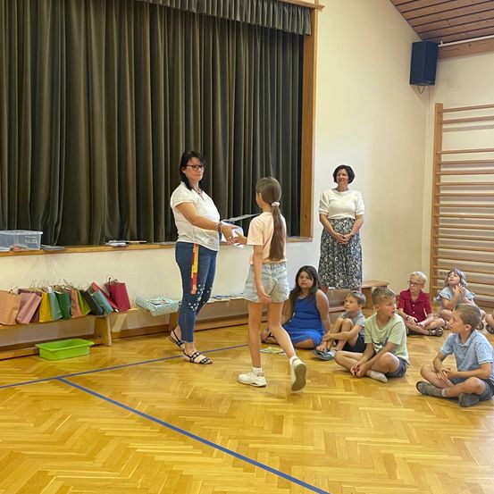 Eine Frau überreicht einer jungen Mädchen ein Zertifikat in einem Raum, in dem Kinder auf dem Boden sitzen. Zwei Frauen stehen hinter ihnen, und ein Lautsprecher ist an der Wand montiert.