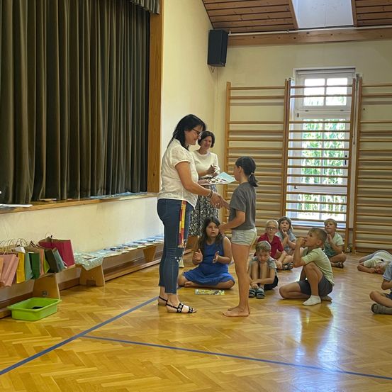 Eine Frau überreicht einer jungen Mädchen ein Zertifikat in einem Klassenzimmer, während andere Kinder auf dem Boden sitzen.
