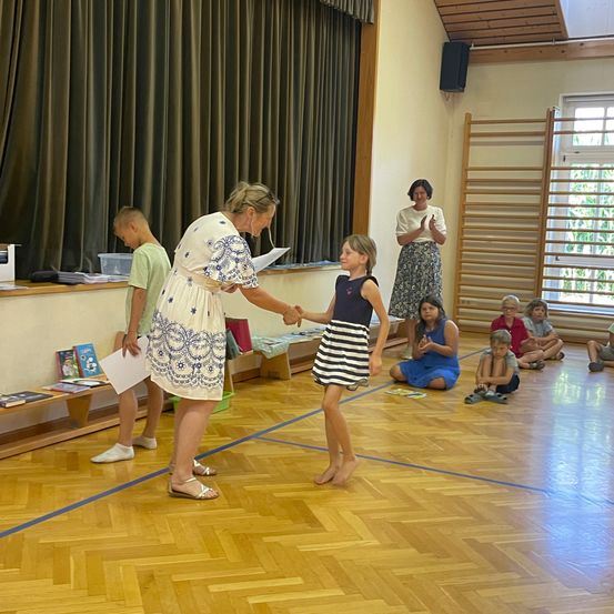 Eine Frau und ein junges Mädchen schütteln sich die Hände in einem Klassenzimmer. Andere Kinder sitzen auf dem Boden und schauen zu. Eine Frau steht hinter ihnen und klatscht.