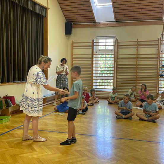 Eine Frau in einem Blumenkleid steht mit einem jungen Jungen und schüttelt ihm die Hand in einem Klassenzimmer, in dem Kinder auf dem Boden sitzen.