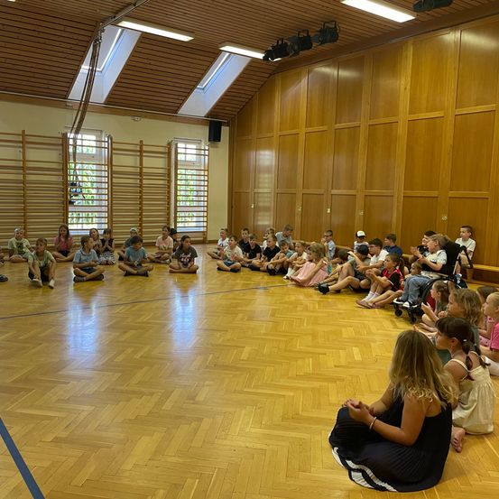 Eine Gruppe von Kindern und Erwachsenen sitzt kreisförmig auf einem Holzboden in einem Raum mit Holzwänden und Fenstern.