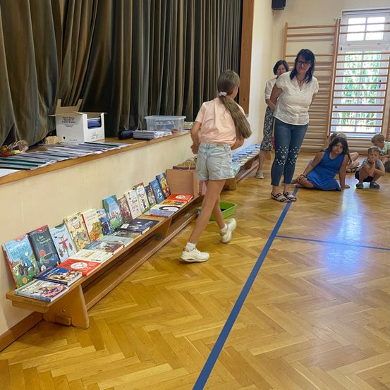 Eine Gruppe von Kindern und Erwachsenen versammelt sich in einem Raum mit einem Holzregal voller Bücher. Eine Frau steht in der Nähe des Regals, und ein Mädchen geht daran vorbei. Andere Kinder sitzen auf dem Boden.